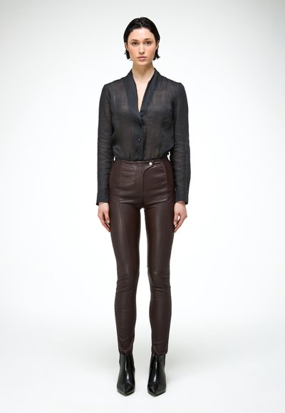 Kobieta stojąca w przezroczystej czarnej koszuli zapinanej na guziki, ciemnobrązowych skórzanych spodniach z wysokim stanem oraz czarnych szpiczastych botkach za kostkę.
