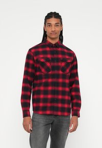 Rød og svart rutete flanellskjorte med lange ermer, to brystlommer og krage med knapper. Brukt med grå jeans.