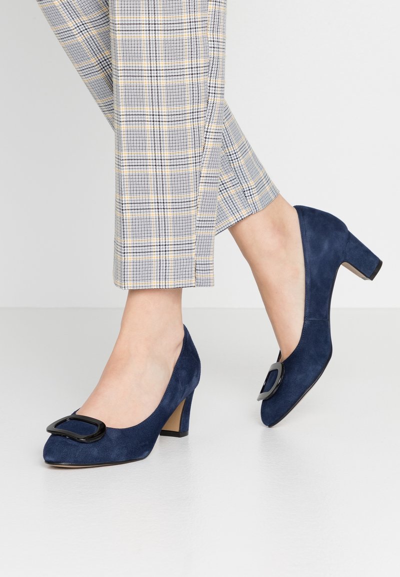 Chaussures en daim bleu marine à talon moyen avec un détail de boucle noire portées avec un pantalon raccourci à carreaux gris sur un fond uni.