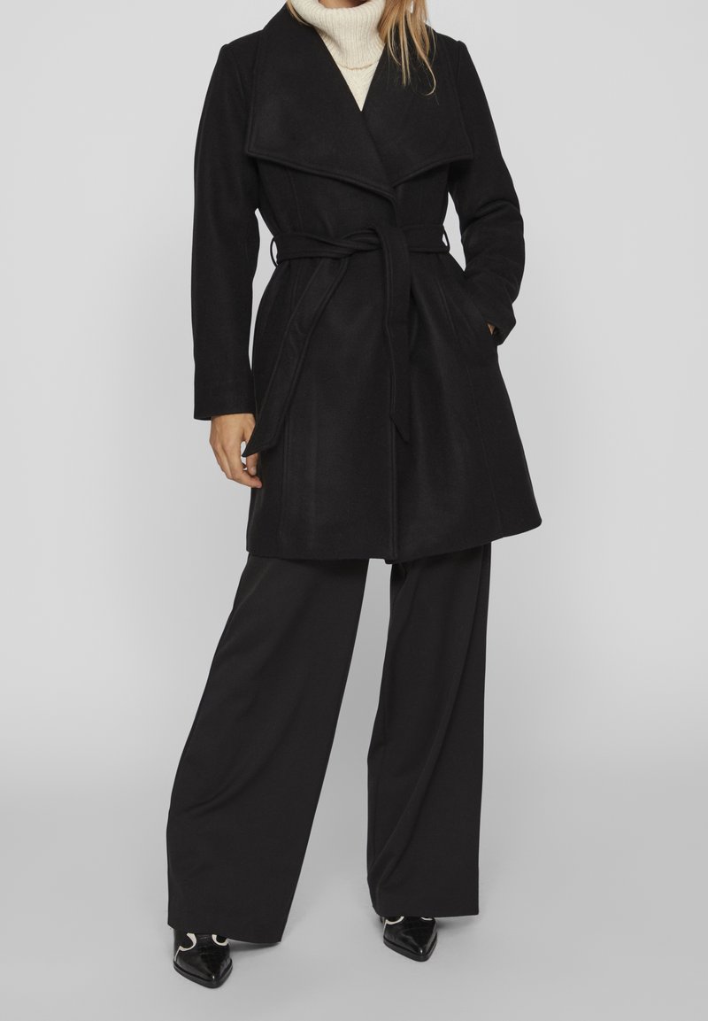 Trench coat nero in misto lana con vita cinturata, ampi revers e vestibilità ampia. Abbinato a pantaloni ampi e scarpe a punta.