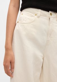 Bílé bavlněné kalhoty s širokými nohavicemi, zdobené zlatými kovovými prvky, kapsami a jemnou texturou. Pas je hladký s klasickým střihem.