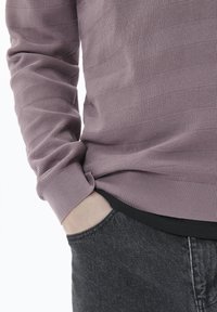 Lila strukturierter Pullover mit horizontalen Streifen, kombiniert mit dunkelblauen Jeans. Das Design betont den Kontrast von Farben und Materialien.