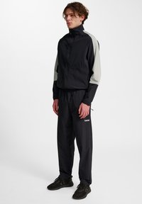 Zwarte jas met grijze mouwen, ritssluiting aan de voorkant en hoge kraag, gecombineerd met zwarte joggers met een klein wit logo. Getextureerde stof.