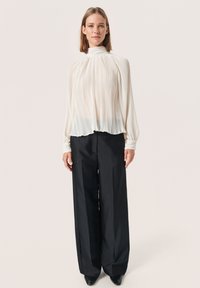 Witte geplooide blouse met een hoge hals en lange mouwen, gecombineerd met zwarte wijde broek en zwarte puntige schoenen. Gladde stofstructuur.