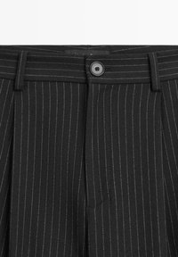 Schwarze Nadelstreifenhose mit Knopfverschluss, ausgestattet mit einer plissierten Vorderseite und strukturierter Stoff. Die Nadelstreifen sind weiß auf schwarzem Hintergrund.