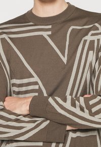 Pull en maille marron avec de grands motifs géométriques abstraits beige clair et col, poignets et ourlet côtelés, porté avec les bras croisés.