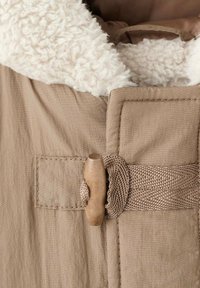 Outerwear beige con un morbido colletto in fleece bianco. Presenta una cintura tessuta e un chiusura a toggles in legno. Tessuto testurizzato con cuciture visibili.