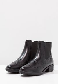 Botas de tobillo de cuero negro con un acabado pulido, paneles laterales elásticos, detalles de brogue y un tacón bajo apilado. Suela de goma texturizada.