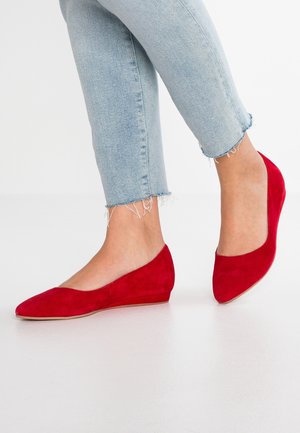 Fødder iført lyse røde loafers i ruskind med spids tå, kombineret med lyseblå jeans med frynset kant.