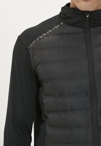Schwarze hybride Jacke mit einem gesteppten Vorderteil und glatten, dehnbaren Ärmeln. Hoher Kragen mit einem Reißverschluss vorne und subtilen Glanzakzenten.