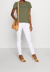 Olivgrön kortärmad skjorta med stjärndekorationer, i kombination med vita slim-fit byxor och ljusbruna sandaletter med öppen tå, bärande en ljusbrun axelremsväska.