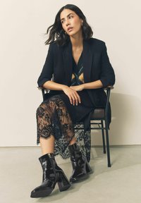 Black patent leather ankle boots with a chunky heel, paired with a black lace dress featuring floral patterns, and a tailored black blazer.