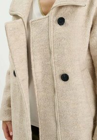 Abrigo de lana beige con una superficie texturizada, grandes botones negros y solapa con cuello; parcialmente desabrochado con una blusa blanca debajo.