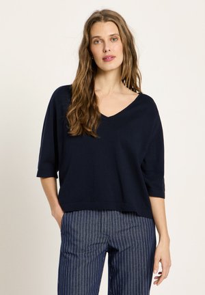 Frau mit langen, welligen Haaren, die ein lockeres, dunkelblaues V-Ausschnitt-Oberteil und blau gestreifte Hose trägt, steht mit einer Hand in der Tasche.