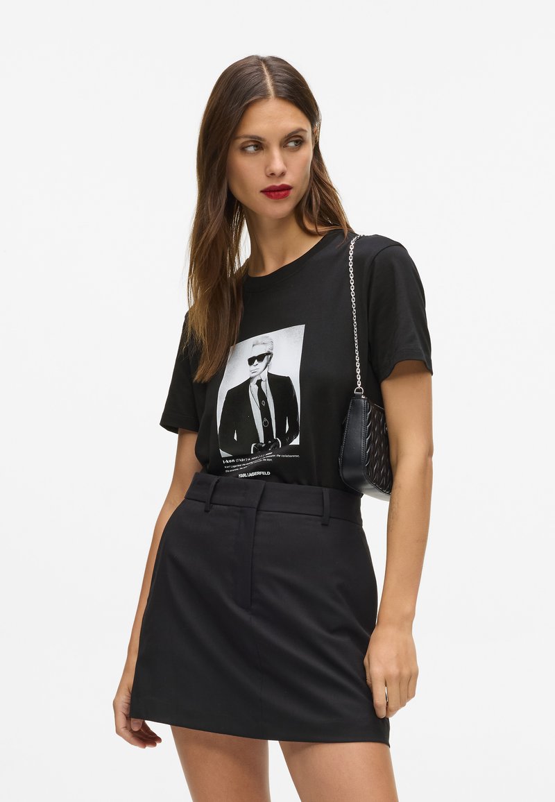 Mustalane puuvillane T-särk, millel on graafiline trükk mees prillides, millele on lisatud kõrge vöökoht must seelik ja väike must käsipak.