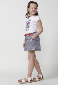 Camicia bianca a maniche corte con stampa grafica, abbinata a una gonna blu navy con motivi, dotata di tasche e una cintura rossa. Indossata con sandali bianchi.