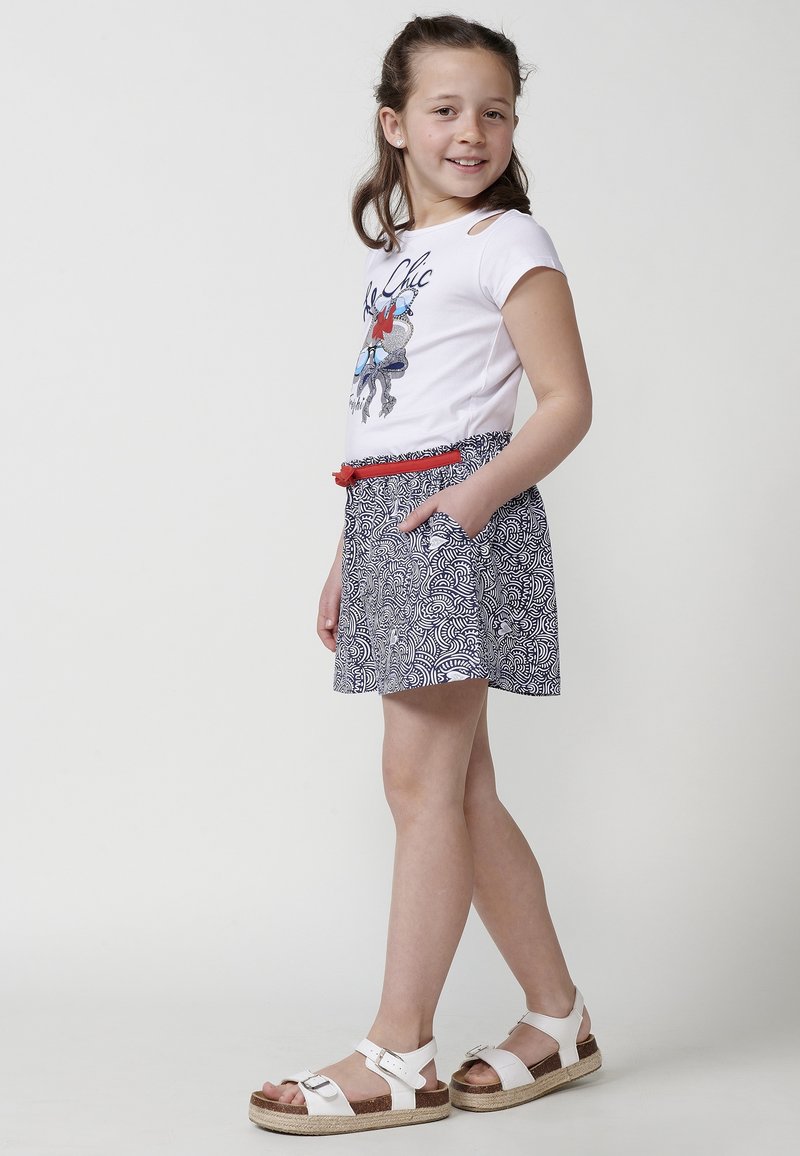 Camicia bianca a maniche corte con stampa grafica, abbinata a una gonna blu navy con motivi, dotata di tasche e una cintura rossa. Indossata con sandali bianchi.