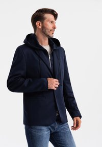 Marine hybrid blazer med hætte, lynlås foran og snore. Blødt stof, glat tekstur og to frontlommer. Afslappet og moderne design.