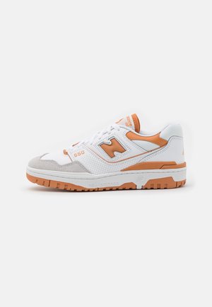 Bílá a oranžová teniska New Balance 550 s šedými semišovými akcenty na špičce a oranžovou podrážkou, zobrazená z boku na bílém pozadí.