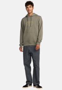 Olijfgroene hoodie met een kangoeroezak, met een gouden logo-print. Gecombineerd met donkergrijze pants en zwarte schoenen.