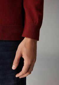 Rød strikket sweater med ribbede manchetter, blød tekstur, båret af en hånd placeret nær mørk denim, der fremhæver ærmedesignet og pasformen.