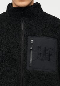 Άνδρας που φοράει μαύρο φλις μπουφάν με φερμουάρ στο μπροστινό μέρος και τσέπη στο στήθος με φερμουάρ, με κεντημένο το λογότυπο "GAP".