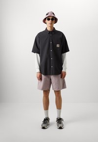 Camicetta nera a maniche corte, textured, con bottoni e logo sulla tasca, abbinata a pantaloni corti rosa pallido, calze bianche, occhiali da sole e un cappello a secchiello rosa chiaro.