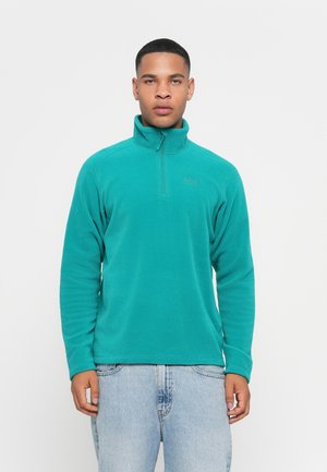 Man in een teal kleurige fleece pullover met kwart rits en lichtblauwe denim jeans, staande tegen een plain witte achtergrond.