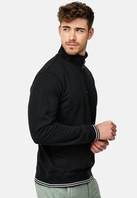 Svart zip-up hettegenser med høy krage, ribbede mansjetter og kant med hvite striper. Ser ut til å være laget av mykt, teksturert stoff.