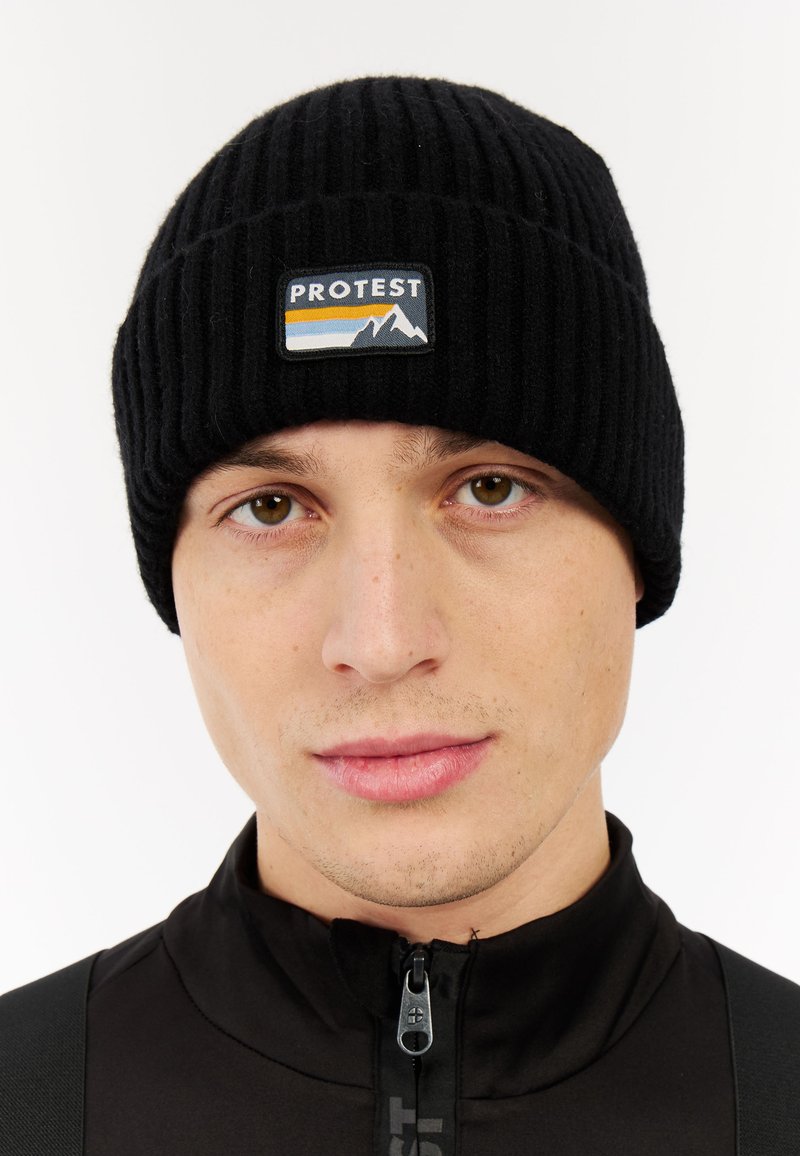 Black ribbed knit beanie with a stitched logo patch featuring blue and yellow stripes above mountains, worn by a person in a black zip-up top.
