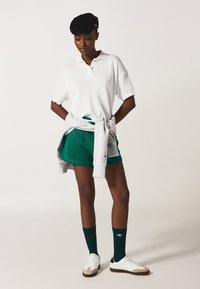 Polo de manga corta blanco, pantalones cortos deportivos verdes, sudadera gris atada a la cintura, calcetines verdes y zapatillas blancas con detalles grises.
