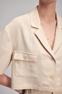 Beige zkrácená bunda s měkkou texturou, krátkými rukávy, klopovým límcem a dvěma knoflíky; obsahuje náprsní kapsu na knoflíkovou sponu.