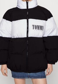 Giacca imbottita in bianco e nero con colletto alto e chiusura con zip. Presenta il logo "TOMMY JEANS" sul petto e polsini e orlo elasticizzati.