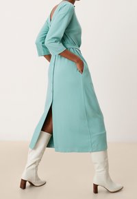 Un abito midi turchese con scollatura a V, maniche a tre quarti, tasche laterali e uno spacco, abbinato a stivali bianchi al ginocchio con tacco.