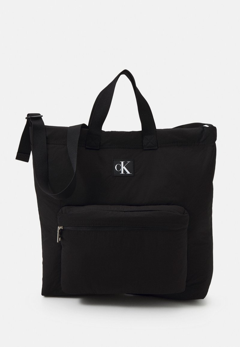 Sac cabas noir Calvin Klein avec doubles poignées, bandoulière réglable et poche zippée à l'avant.