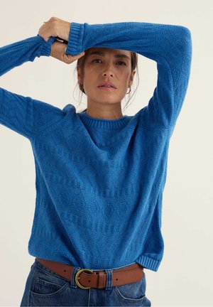 Femme portant un pull bleu texturé et une ceinture marron avec une boucle en or, levant les bras au-dessus de la tête sur un fond uni.