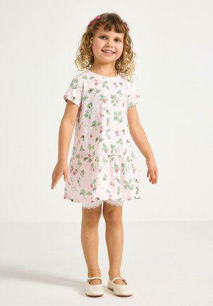 Smilende ung pige med krøllet hår iført lyserød blomsterkjole med blonder på kanten, shorts og hvide sko, stående mod en ensfarvet baggrund.