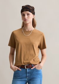 Braunes Baumwoll-T-Shirt mit Rundhalsausschnitt, kombiniert mit blauen Jeans und einem braunen Gürtel. Das Model trägt einen gemusterten schwarzen Schal und eine silberne Halskette.