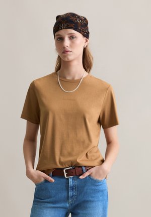 Brown cotton t-shirt with a crew neck, paired with blue denim jeans and a brown belt. Model wears a patterned black scarf and silver necklace.