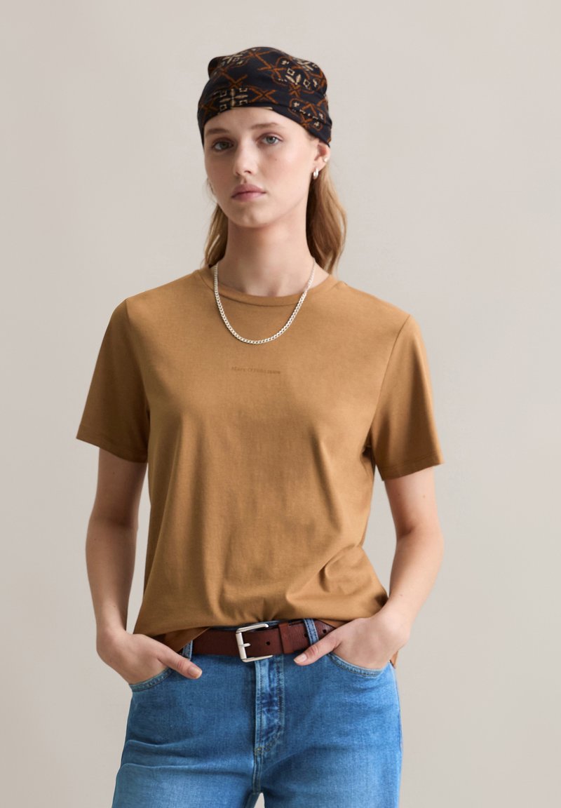 Braunes Baumwoll-T-Shirt mit Rundhalsausschnitt, kombiniert mit blauen Jeans und einem braunen Gürtel. Das Model trägt einen gemusterten schwarzen Schal und eine silberne Halskette.