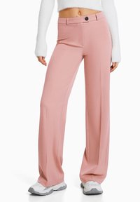Světle růžové kalhoty s širokými nohavicemi, hladkým povrchem, předními kapsami a zapínáním na knoflík v pase, doplněné bílými teniskami.