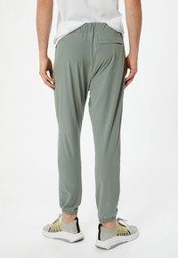 Lichtgroene, elastische broek met taillesluiting en manchetten bij de enkels. Glad materiaal en een enkele achterzak. Gepaarde met grijze en gele sneakers.