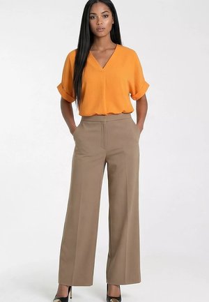 Kobieta stojąca z rękami w kieszeniach, ubrana w pomarańczową bluzkę z krótkim rękawem, beżowe szerokie spodnie i czarne szpilki na białym tle.