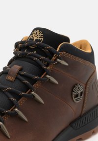 Bota de senderismo de cuero marrón con un cuello de malla negra, logo dorado y ojetes de metal. Presenta una superficie texturizada y acentos de costura naranjas.