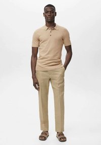 Polo beige con maniche corte, colletto a tre bottoni e pantaloni khaki. I sandali hanno molteplici cinghie e una suola piatta.