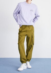 Jasnofioletowa bluza z logo na piersi, w parze z oliwkowymi spodniami typu cargo z kieszeniami i elastycznymi mankietami przy kostkach. Białe trampki.