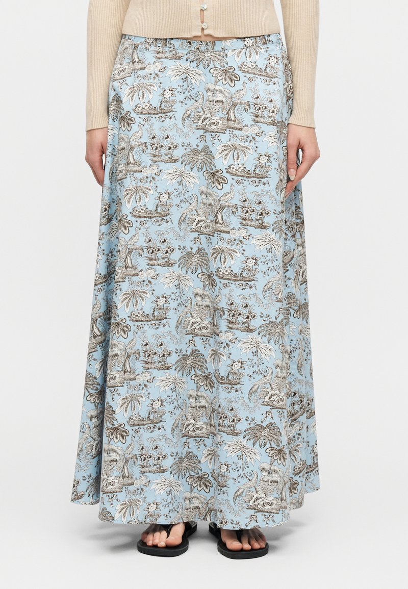 Γυναίκα που φοράει μπεζ μακρυμάνικο τοπ και μακριά φούστα με μπλε λουλουδάτο σχέδιο, με μαύρα σανδάλια ανοιχτά στα δάχτυλα, στέκεται μπροστά σε λευκό φόντο.