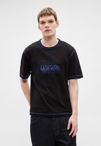 T-shirt preta em algodão com detalhes de costura azul. Apresenta uma estampa gráfica azul com o texto "HUGO" ao centro. Gola redonda.