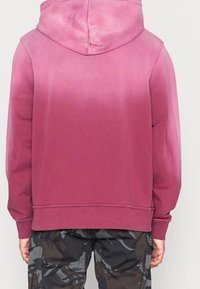 Mikina s kapucí má gradientní design v odstínech růžové, je vyrobena z jemné bavlny, má žebrované manžety a lem, a je kombinována s maskáčovými kalhotami.