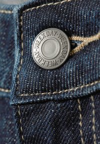 Donkerblauwe denim stof met een close-up van een metalen knop met de inscriptie "WEEKDAY." Bevat zichtbare steken en de textuur van de stof.